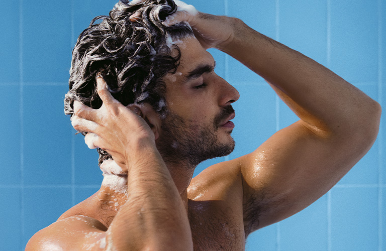 Foto de un hombre en la ducha lavándose el pelo con espuma. Fondo de azulejos azules. Se ven los brazos levantados, el torso mojado y las manos masajeando el cuero cabelludo.